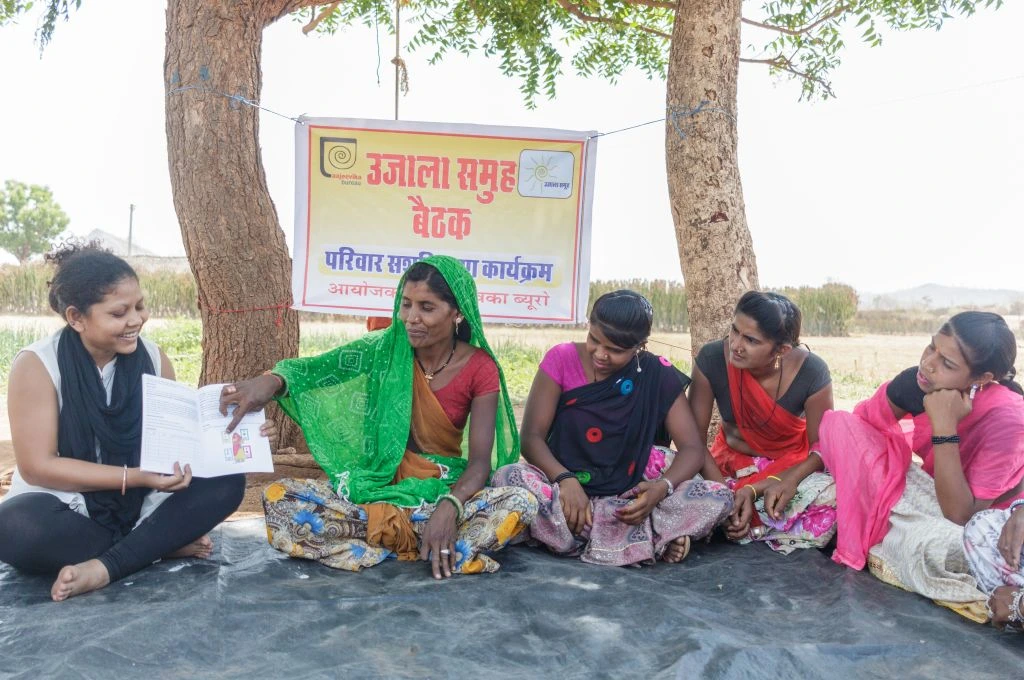 The image shows a group of women sitting under the shade of trees in an open ground in a rural area. There is a banner hanging from the trees which reads 'Ujala Samooh Baithak' or a meeting of the Ujala group. One of the women, seemingly not from the village, is holding a book in her hands as one of the women from the village points to a diagram on the open page.