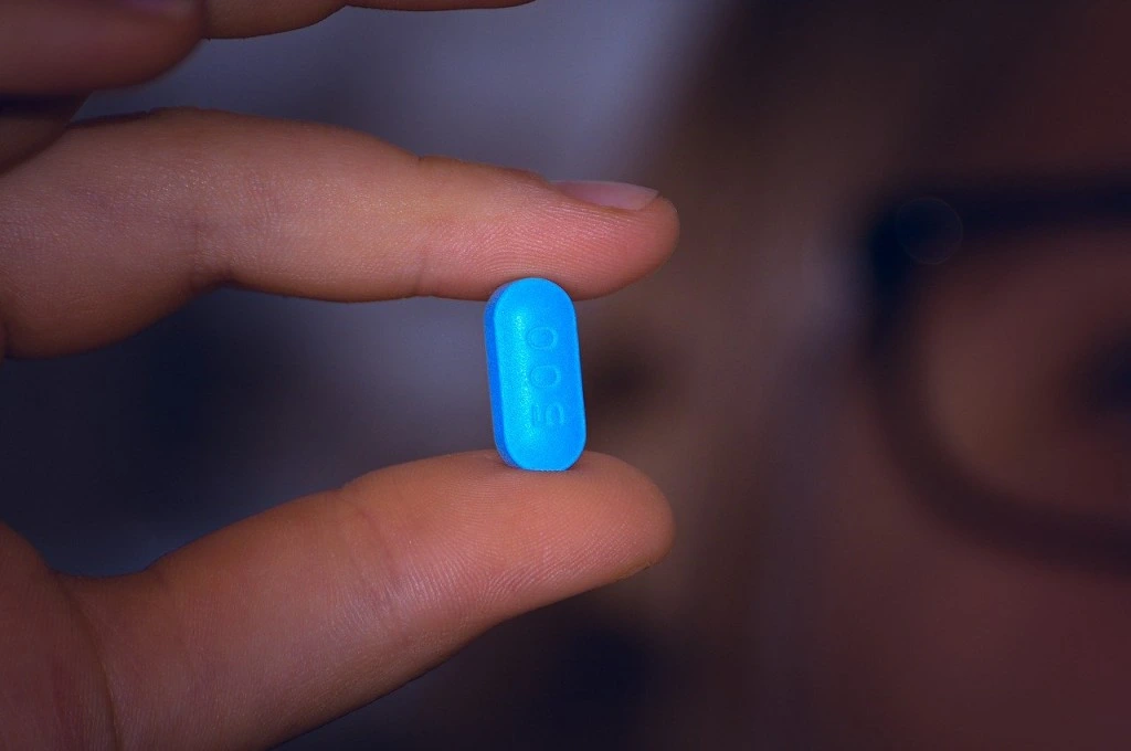 The image is a close-up shot of a person's hands and they are holding a blue medicine tablet using their thumb and forefinger.