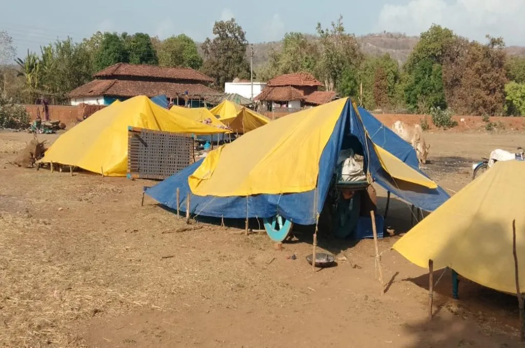 The image shows a few temporary shelters made from tarpaulin and bamboo sticks in an open ground. People's belongings are visible through the gaps in the sheets of tarpaulin. There is a building in the distance in the background.