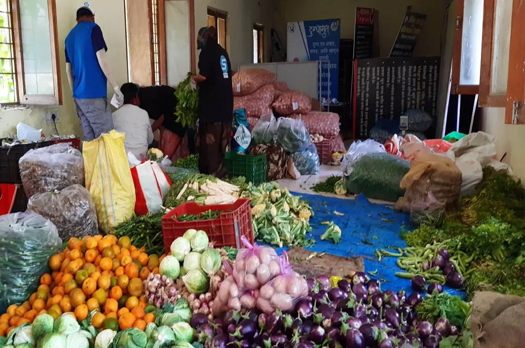 The image is set inside the room of a building. Across the floor, there are piles of different vegetables including onions, brinjals, cabbage, and cauliflower. Onions have also been packaged into burlap sacks. There is also a pile of oranges and three people are working together in a corner. There are various informational hoardings and posters stored at the other end of the room.