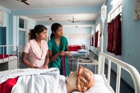 Two young girls in a hospital standing next to a hospital bed with a manequin-skill development programmes