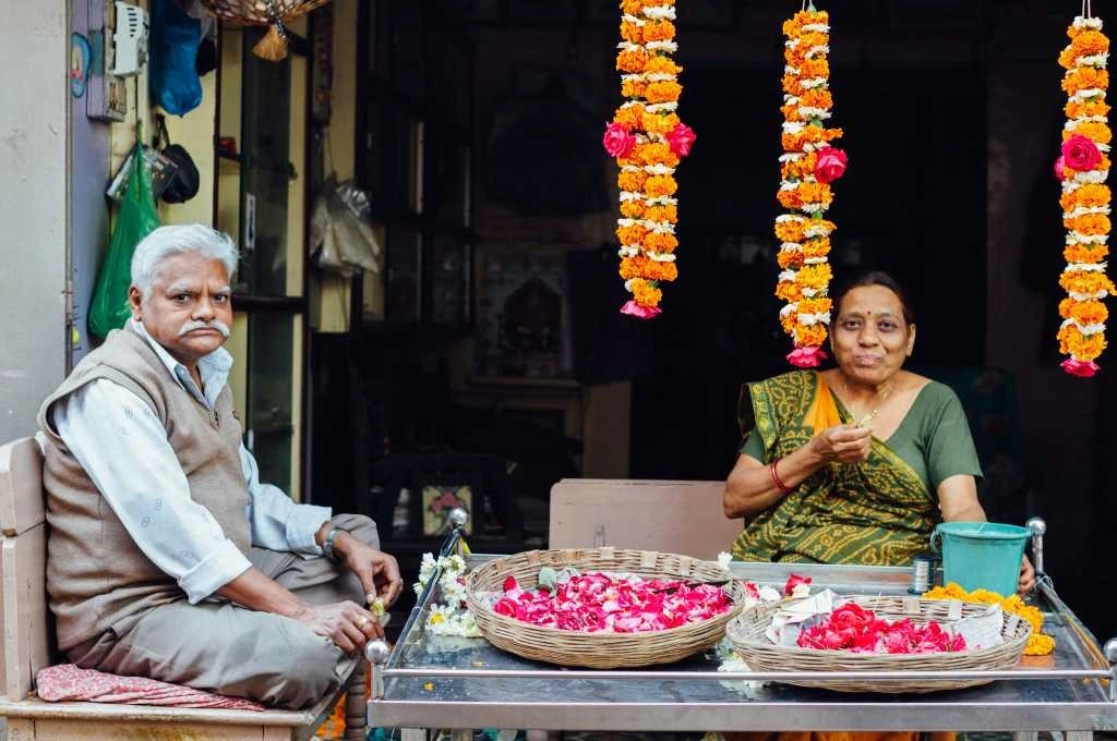 The image shows a middle-aged man and woman sitting at a table outside a shop. There are garlands strung from the ceiling and there are baskets of rose petals placed on the table._Sarkari loan COVID-19