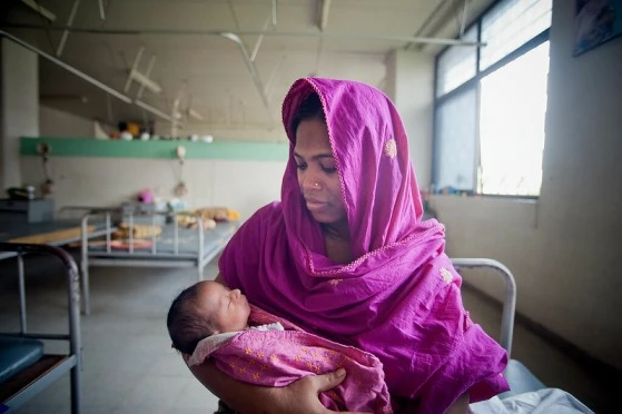 The image shows a woman sitting in a hospital ward. She is holding a baby in her arms._Covid-19 pregnant women