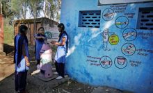 three girls standing around an incinerator as the girl in the middle disposes a menstrual health product into the incinerator. There is a blue wall to their right with wall art about how to safely use a sanitary napkin-menstrual health and hygiene COVID-19
