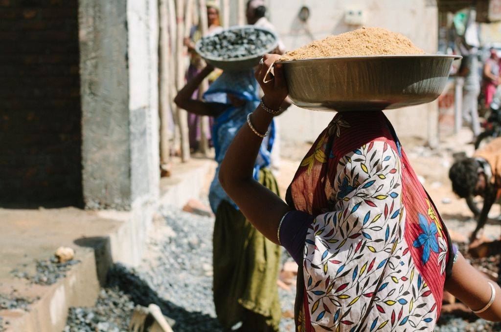 Two women in sarees, carrying construction materials on their head. The photograph has been clicked from behind. NREGA is crucial to the rural economy of India, especially in 2021 with low yield and poor monsoon