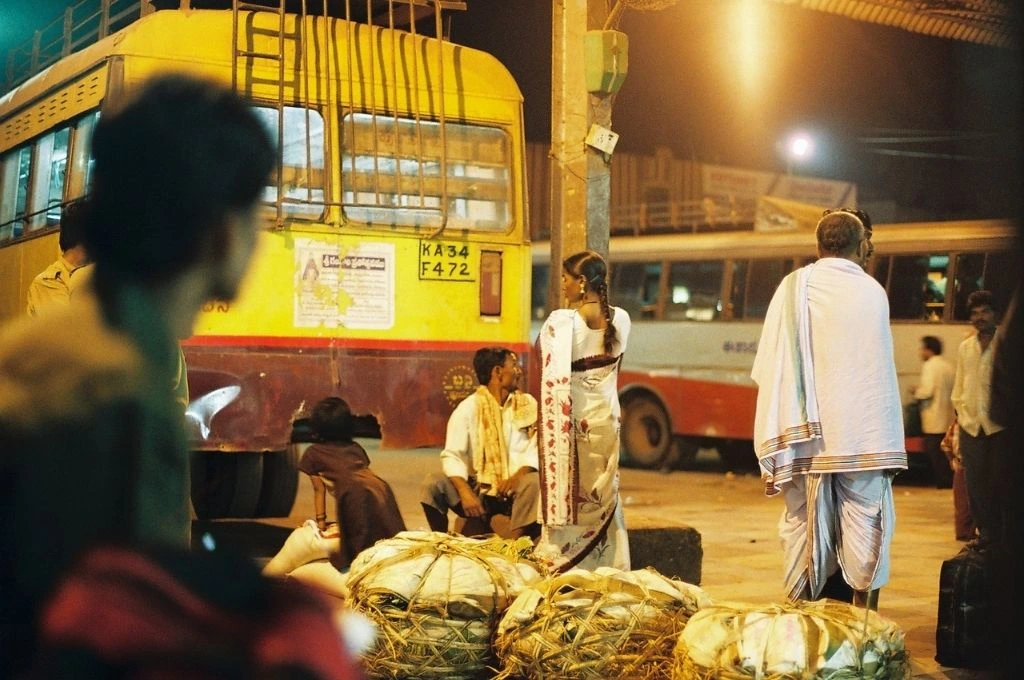 The image shows a bus stop at night. There are two buses parked in the background. There are men and women sitting and standing in the area with their luggage and belongings._Women safety