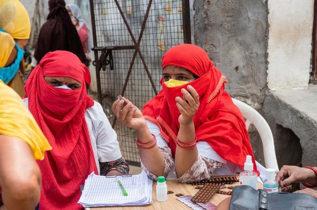 Two Ashas, their face covered with red dupattas, giving medicines to a person. Community healthcare workers were crucial to India’s COVID-19 response. They need better pay and training for their well-being.