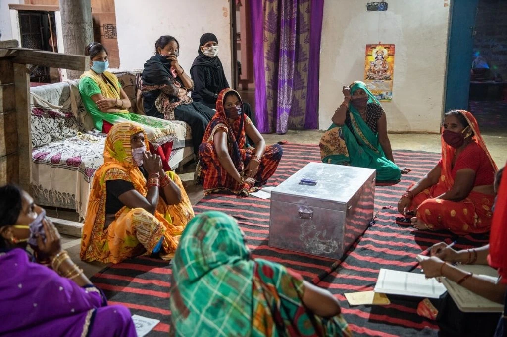 The image features a group of women in a room inside a house in a rural area. Most of the women are sitting in a circle on a carpet on the floor. In the middle, there is a rectangular trunk made of steel. Three women are sitting on a sofa in the background._Women self help group