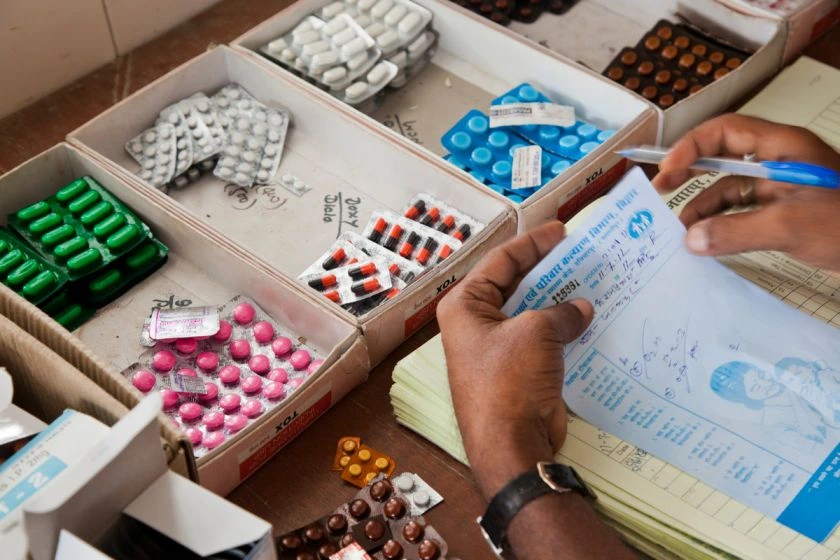The image shows three carboard boxes placed on a table each with strips of different medicine tablets arranged in rows. There are a few strips on the table and a person's hands can be seen as they hold a receipt and a pen._Caste