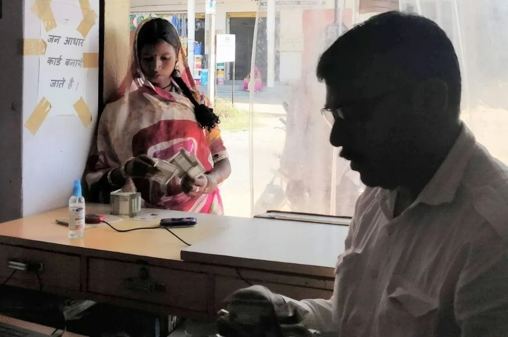 The image shows a young woman standing at the counter of a small shop in a rural area. She is counting money. The sign on the wall next to her reads "Aadhaar cards are made here". A man sits across the counter inside the shop._Banking fraud