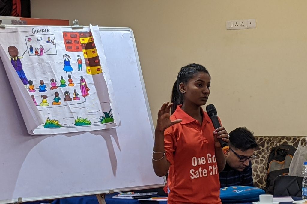 A girl in a red tshirt with a mic speaking to an audience. There's a white board in the background. Lusi Sharma trains and conducts workshops on gender equality