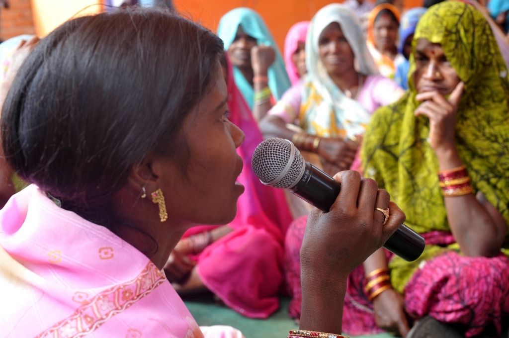 A woman with a mic in a pink saree talking to a gathering of women