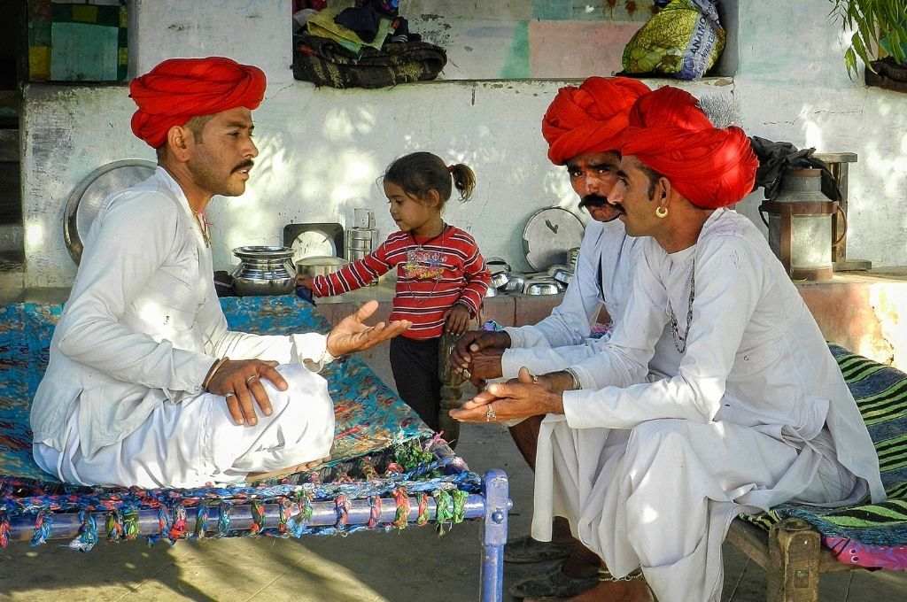 Three men wearing red turban and white kurta pajama sitting on charpoys with a young girl in the background