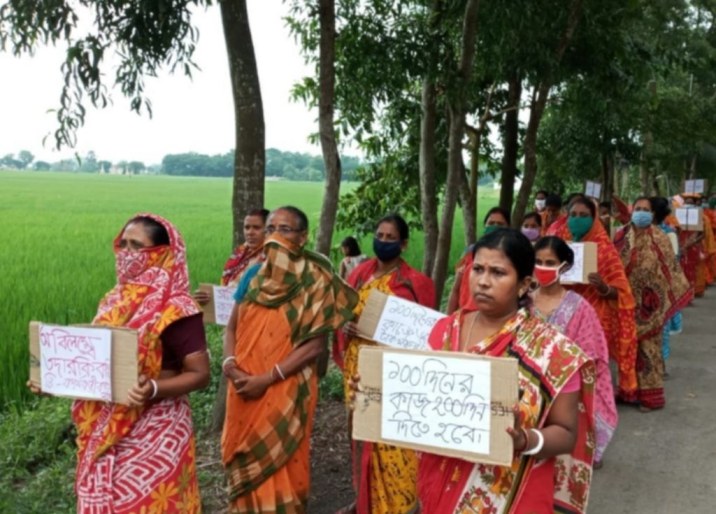 A group of sari-clad women on a morcha. They are holding signs and walking down a road surrounded by green fields_Paschimbanga Khetmajdoor Samity-Budget 2022 MGNREGA