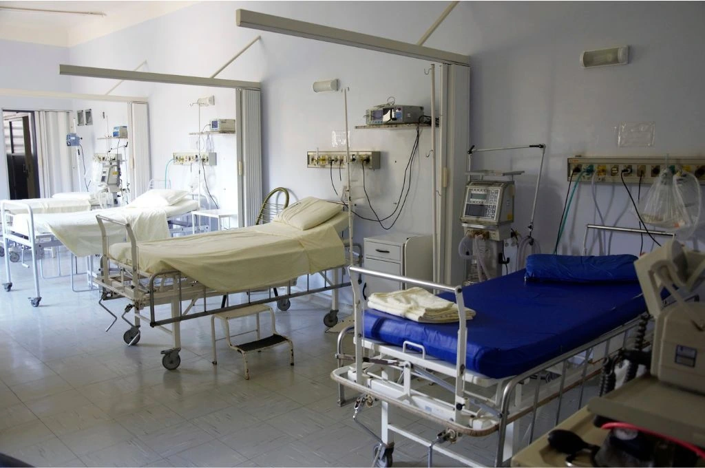 The image shows a hospital ward. There are no people in the ward but it has four hospital beds, each with monitoring machines and other medical objects._Adivasi