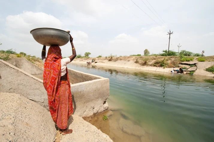 woman carrying a vessel on her head standing near a canal-climate change