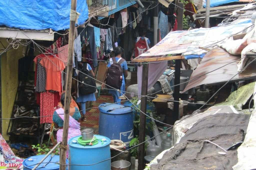 The image is a close-up photograph of an urban informal settlement. A narrow lane passes between two houses. There are big plastic water barrels and other household items placed in front of the houses and clothes have been hung to dry along their exterior. Girls in school uniforms walk down the lane._Adolescent girls