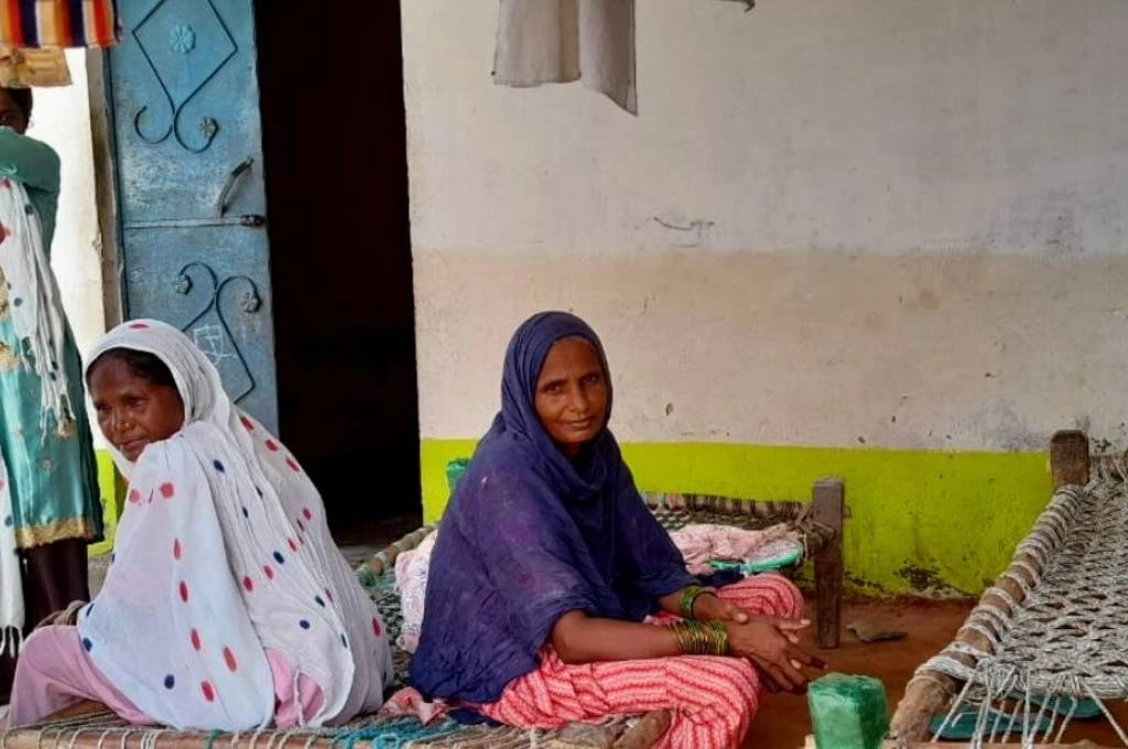 The image features two middle-aged women sitting on a woven bed with a wooden frame. They are seated in a courtyard-like area typically found in rural homes._Electricity