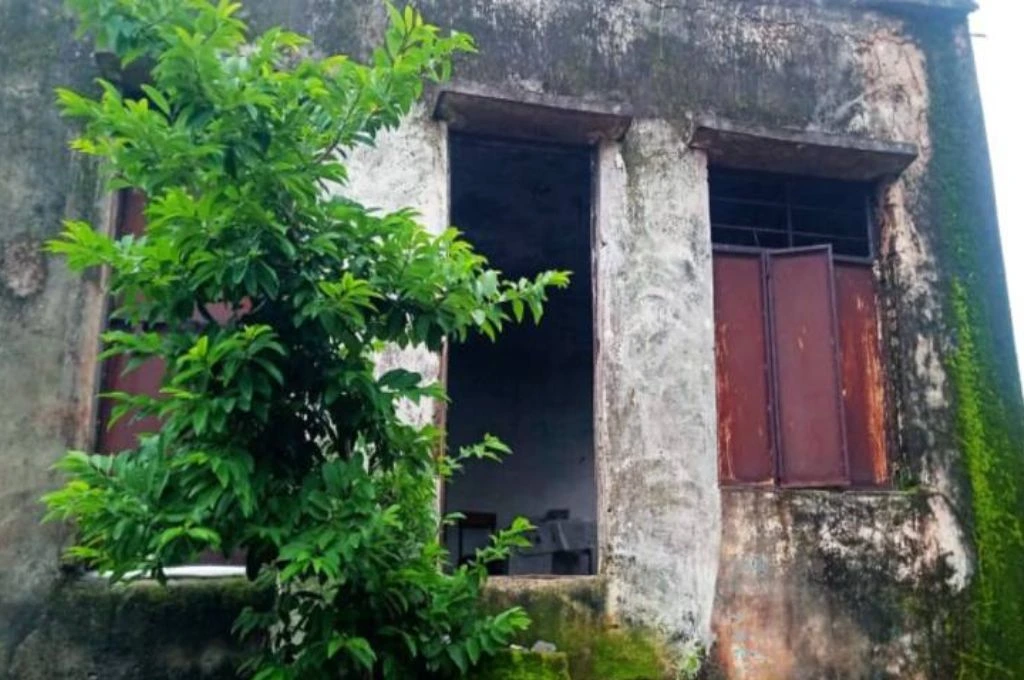 The image shows the exterior of a small building. The metal doors of the building have rust on them and paint on the walls is old and blackened in places._Anganwadi