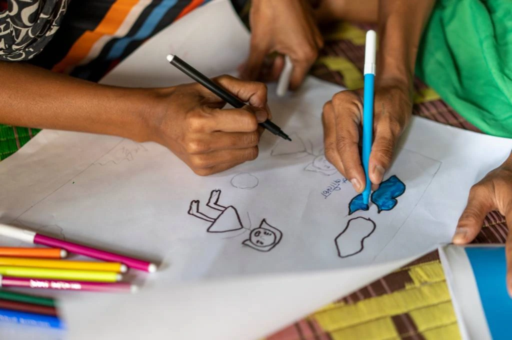 The image is a close-up of two people's hands as they draw on a piece of chart paper. One person uses a blue sketch-pen to colour in clouds while the other draws human figures._school