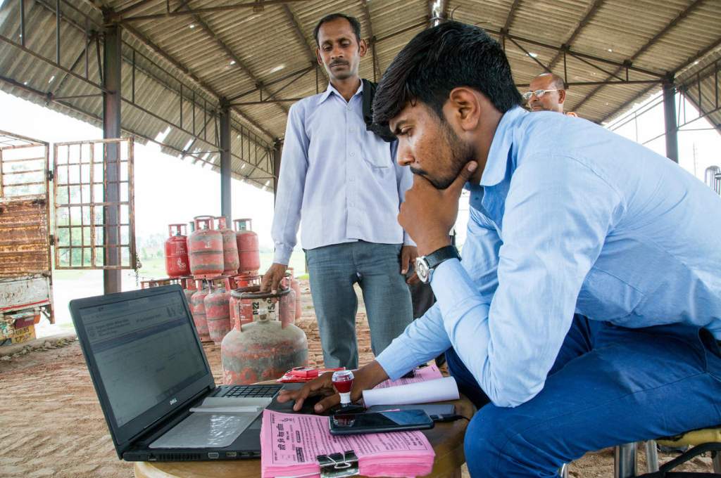 A man using laptop under a shade while another man waits in line with a cylinder_technology for nonprofits