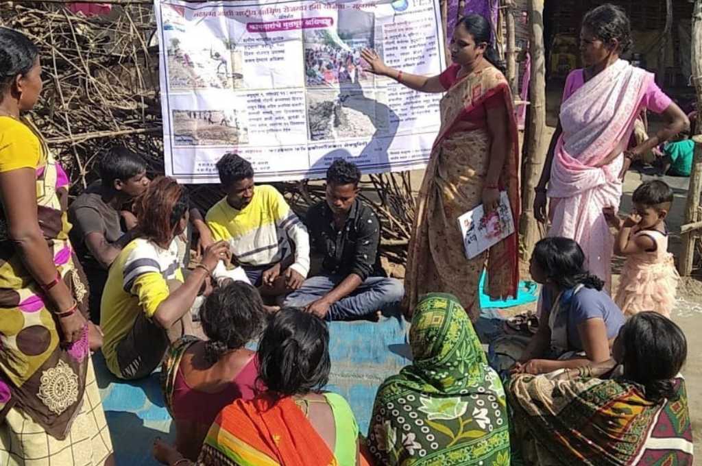 A woman pointing at a poster and speaking. A group of men and women sitting on the ground and looking at her_sustainable agriculture