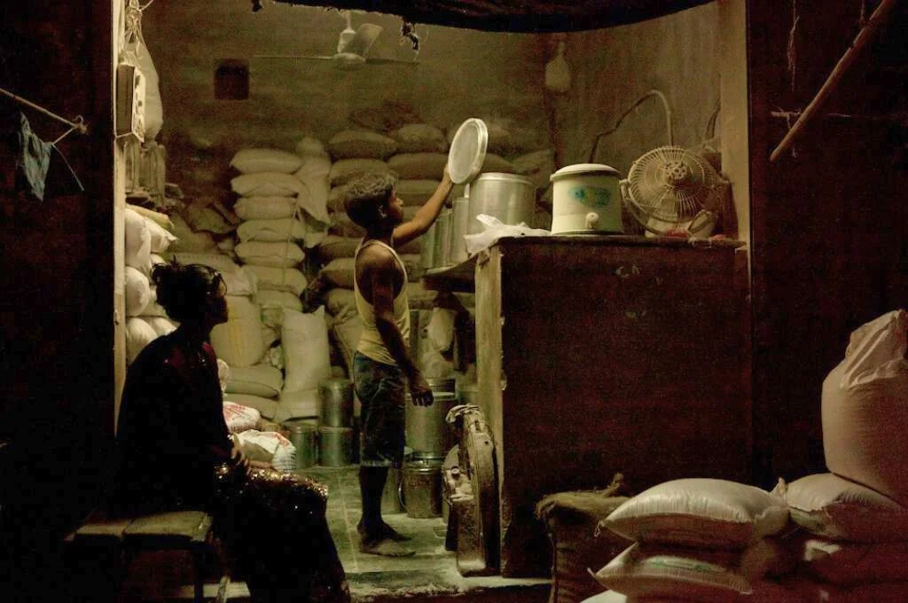 a boy holds the lid of a tin as a woman looks at him, with sacks in the background--livelihoods