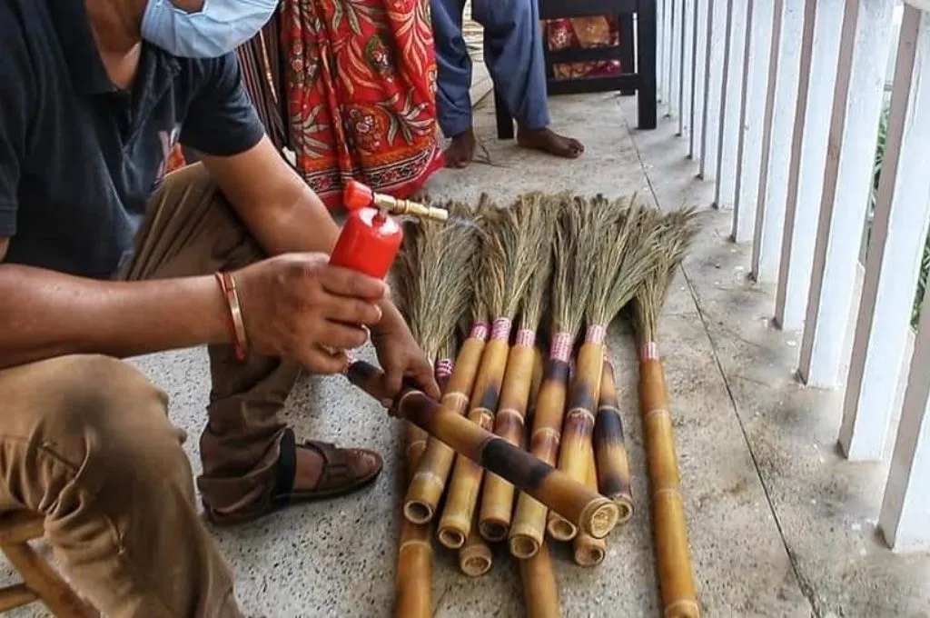 A person uses a small blowtorch to prepare a bamboo stem to make brooms while a bundle of finished brooms made of bamboo stems and grass lay on the ground- Bamboo products