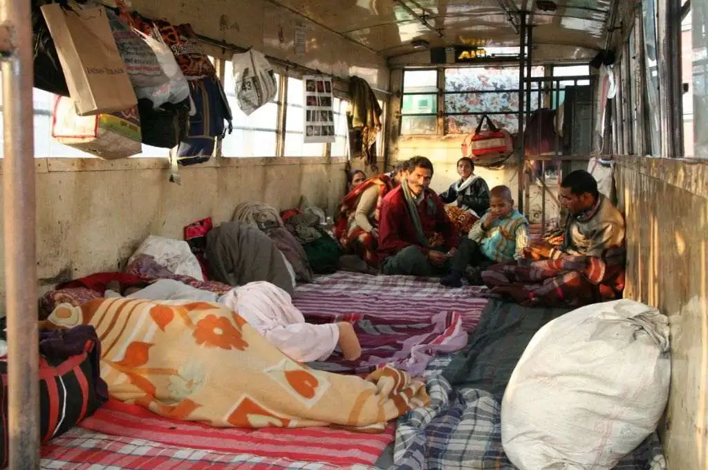 The image shows at least eight people inside an old bus that has been converted into a makeshift shelter. Blankets cover the floor and people's belongings, including bags, are stored on the floor and hung from the windows. Two people are shown sleeping on the ground, while a group of people sit together in the background. - Homeless