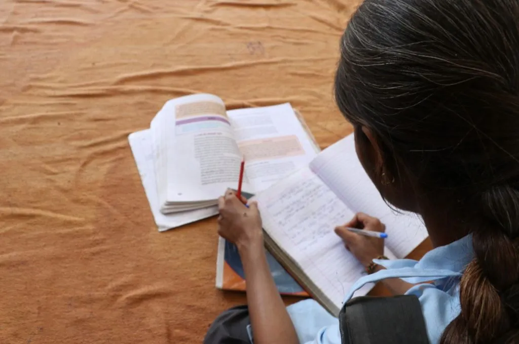 An adolescent girl wearing a school uniform sits with her back to the camera. She reads from a textbook lying open in front of her and writes down her notes in a notebook.- intergenerational change