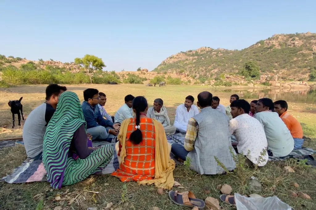 A group of 15 men and women hold a meeting of the tank management committee in the Baura village. They sit in a cirlce in an open area while animals graze on the grass in the background- Water management