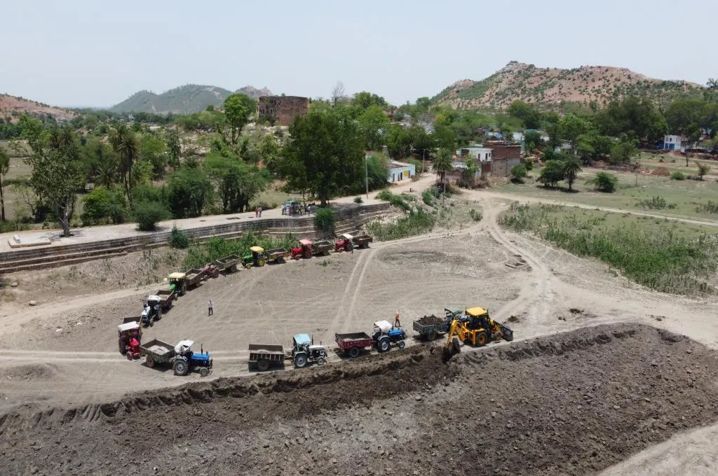 Tractors with trolleys line up to collect silt from a JCB excavator and remove it from the Kudar tank site - Water management