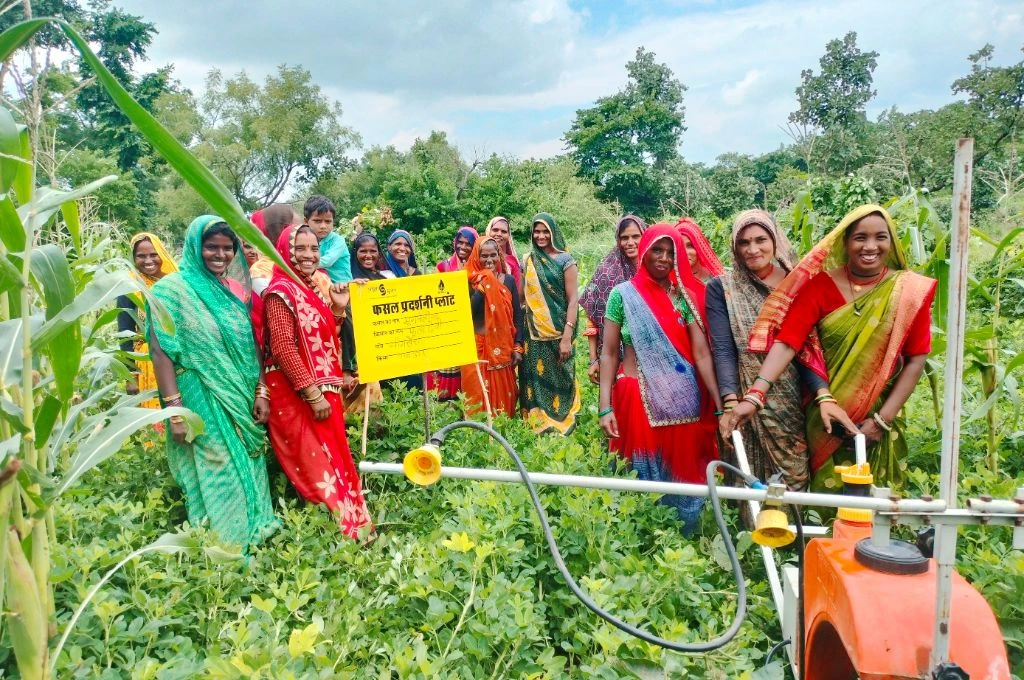 A group of women pose with a sign labelled Crop Exhibition Plot in the background as two women hold onto a sprayer machine- Water management