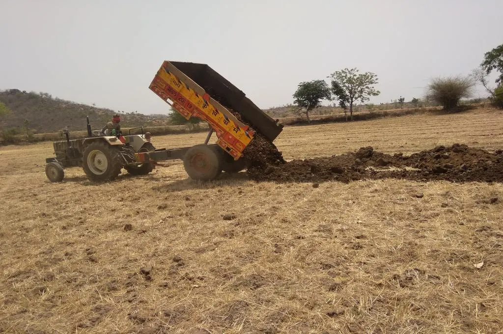 A farmer empties a tractor-trolley full of silt onto his field. There are no crops planted and the field is covered in dried grass- Water management