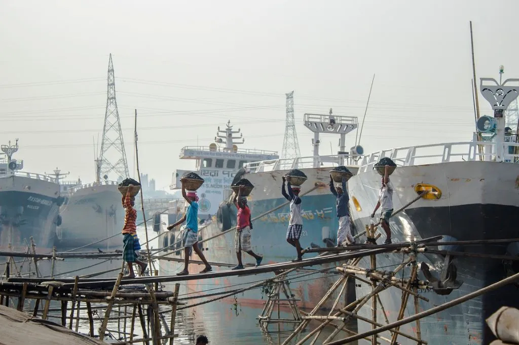 Workers walking on a makeshift bridge carrying baskets of material on their heads from a ship onto a port. - Bonded labour