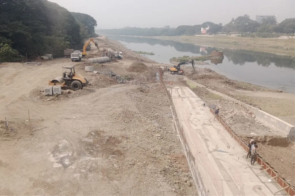 The image shows ongoing construction work on the bank of the Mula-Mutha river. The land has been cleared of any grass and it is now just loose soil. A road-roller stands parked and there are two cranes and a truck with a cement mixer attached to it. A few workers are building a concrete path._Riverfront development