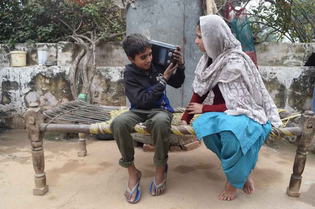 A boy and a woman listen to a radio on a charpai in a rural courtyard_community radio