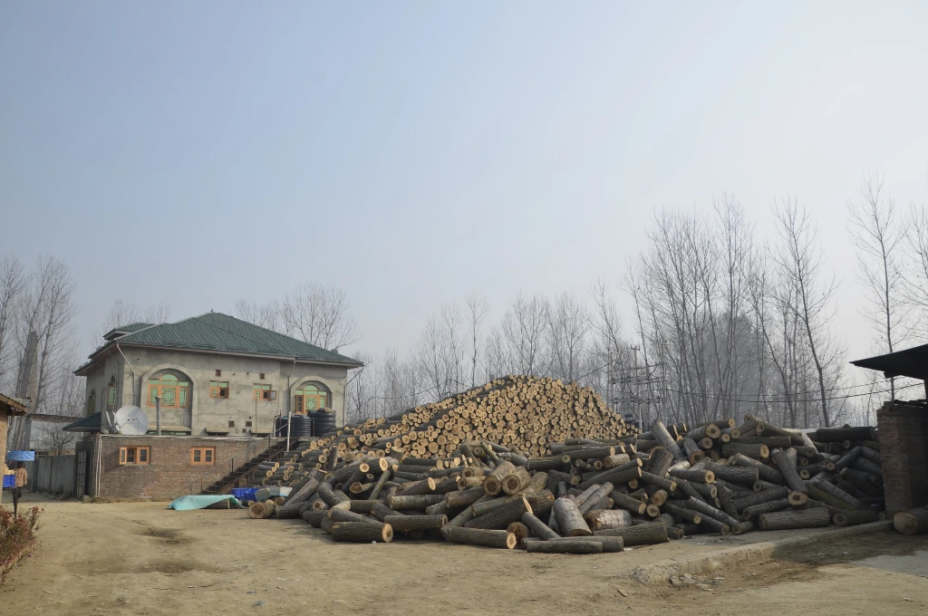 The image features a pile of chopped wooden logs piled in the open. Behind the pile, logs have been stacked in neat rows on top of each other. In the background, there is a large residential structure with bare trees on either side._Oukhoo pencil village