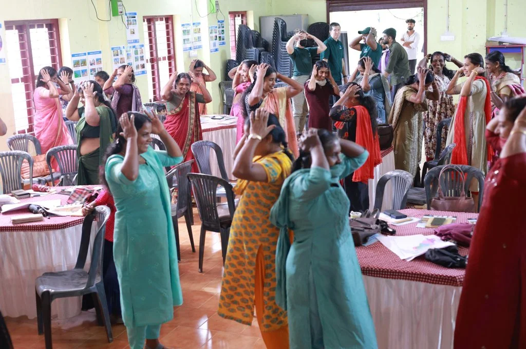 The image depicts a group of women in a room. They are divided into small groups, each with their own table and chairs. The smaller groups of women stand next to their tables with both hands placed on their heads. They are all smiling._Disaster management