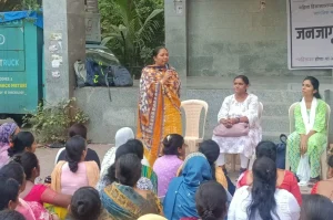 A woman in a yellow and orange saree speaks into a microphone during a community meeting attended by a group of women seated in front of her. Two other women, dressed in white and green, are seated beside her on plastic chairs. The gathering is taking place in an outdoor public space with a stone backdrop and a banner in Marathi_domestic violence
