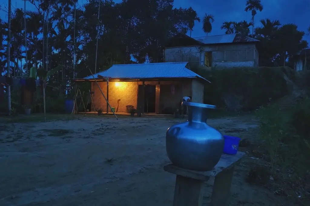 a hut at dusk with a yellow bulb lit on the porch. There are trees in the background and a second hut built on slightly elevated ground.--bamboo-based livelihoods