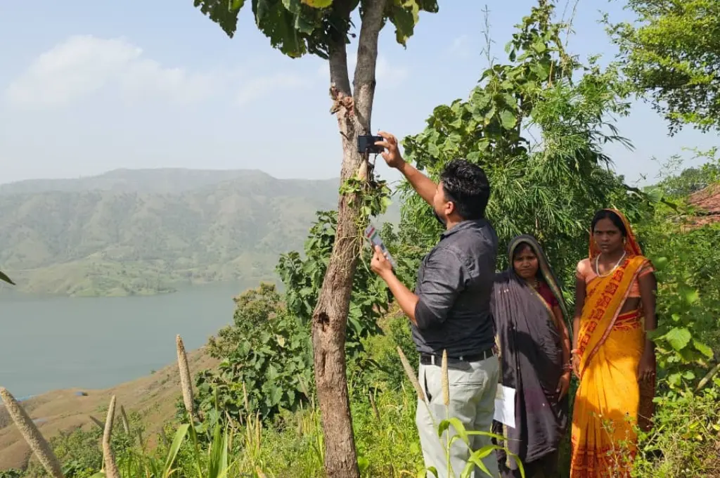two women look into the camera as a man standing next to them with his back towards the camera holds up a phone while standing next to a tree. In his other hand is an Aadhaar card. There is a lake and hills in the background--e-KYC