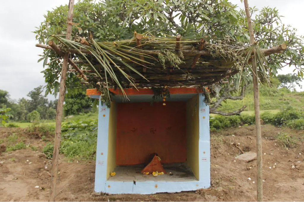 The image shows a small temple-like structure made of concrete. There is a thatched roof made of leaves covering the temple. Inside, there are some religious motifs painted on the wall, with a small pyramid-like structure placed on the ground in the centre of the temple. In the background, there are trees and grass. --Lambani community