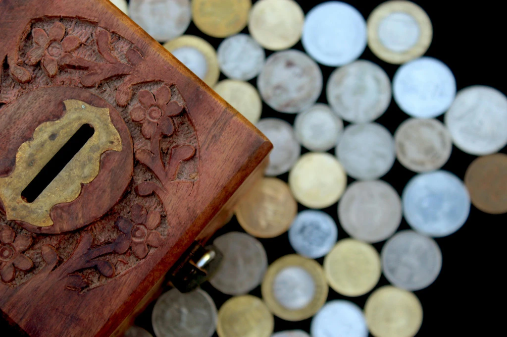 close-up of a carved wooden money box with a brass coin slot, positioned next to an assortment of scattered coins on a dark surface--crowdfunding