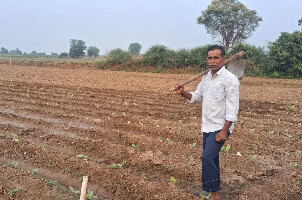Udaysingh Makwana stands in a cultivated farm field holding a hoe over his shoulder, with neat rows of young plants in the soil and trees visible in the background under an open sky.--social sector