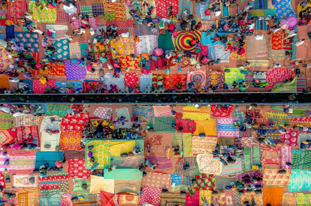 An aerial view of a crowded outdoor market where vendors sit on colorful patterned blankets arranged in rows on both sides of a narrow walkway. People move between the blankets, creating a mosaic-like scene filled with bright fabrics, patterns, and bustling activity--collaboration