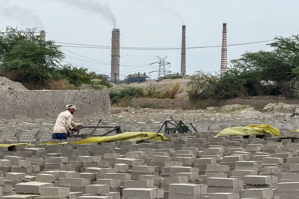 A worker arranges fly ash bricks with smoke coming out of chimneys in the background--fly ash