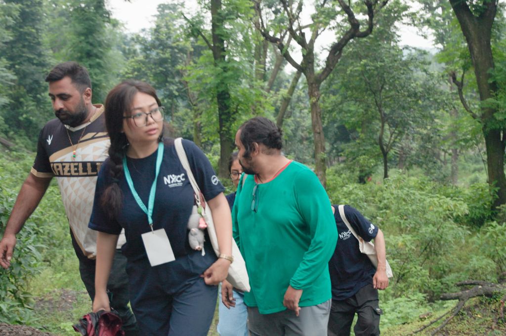 Ishika walks along a narrow forest trail with a small group during an outdoor activity. She carries a shoulder bag and looks to the side as tall trees, dense greenery, and uneven ground surround the group in a lush woodland setting--fellowships