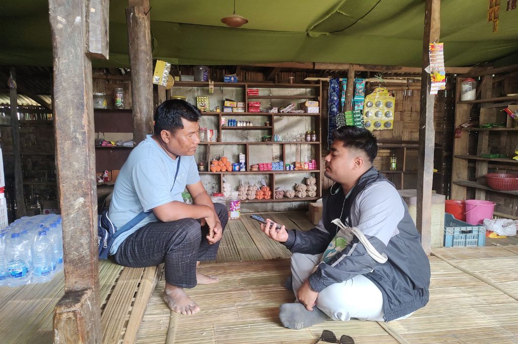 Anupam and another man sit on a bamboo floor inside a small rural shop, engaged in conversation during an interview. Anupam holds a smartphone as a recorder while the other listens attentively. Shelves behind them display packaged snacks, household goods, and bottled water, highlighting a local village store setting--fellowships