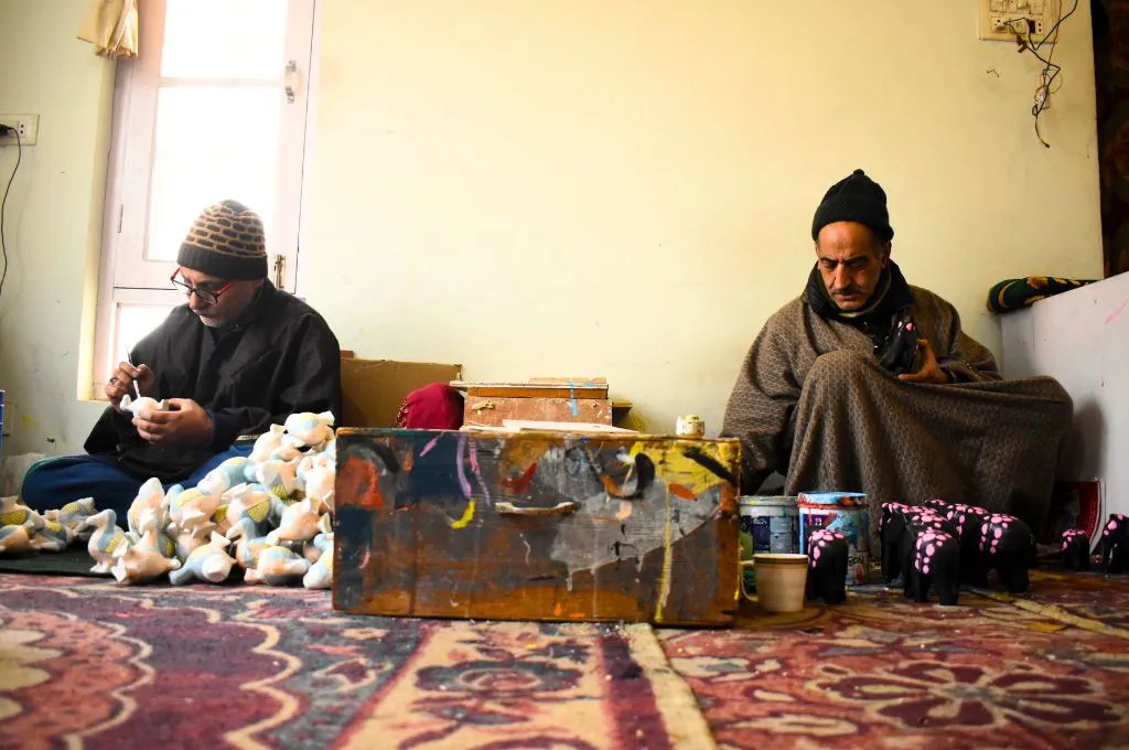 two paper-mache artisans sitting on the carpeted floor of a workshop, with ornaments and paint laid before them--paper-mache in Kashmir